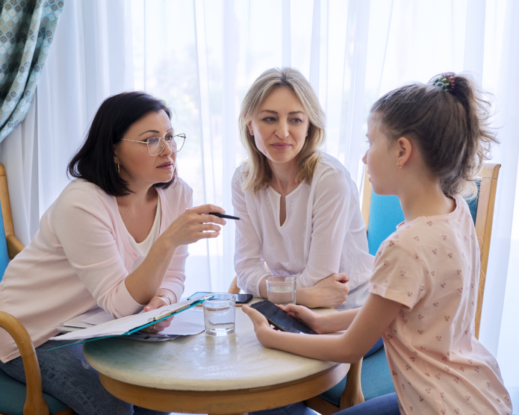 Parent attending therapy with a child. The therapist is at the table with them, pointing her pen at the little girl and has a clipboard in her hand while the mom watches.