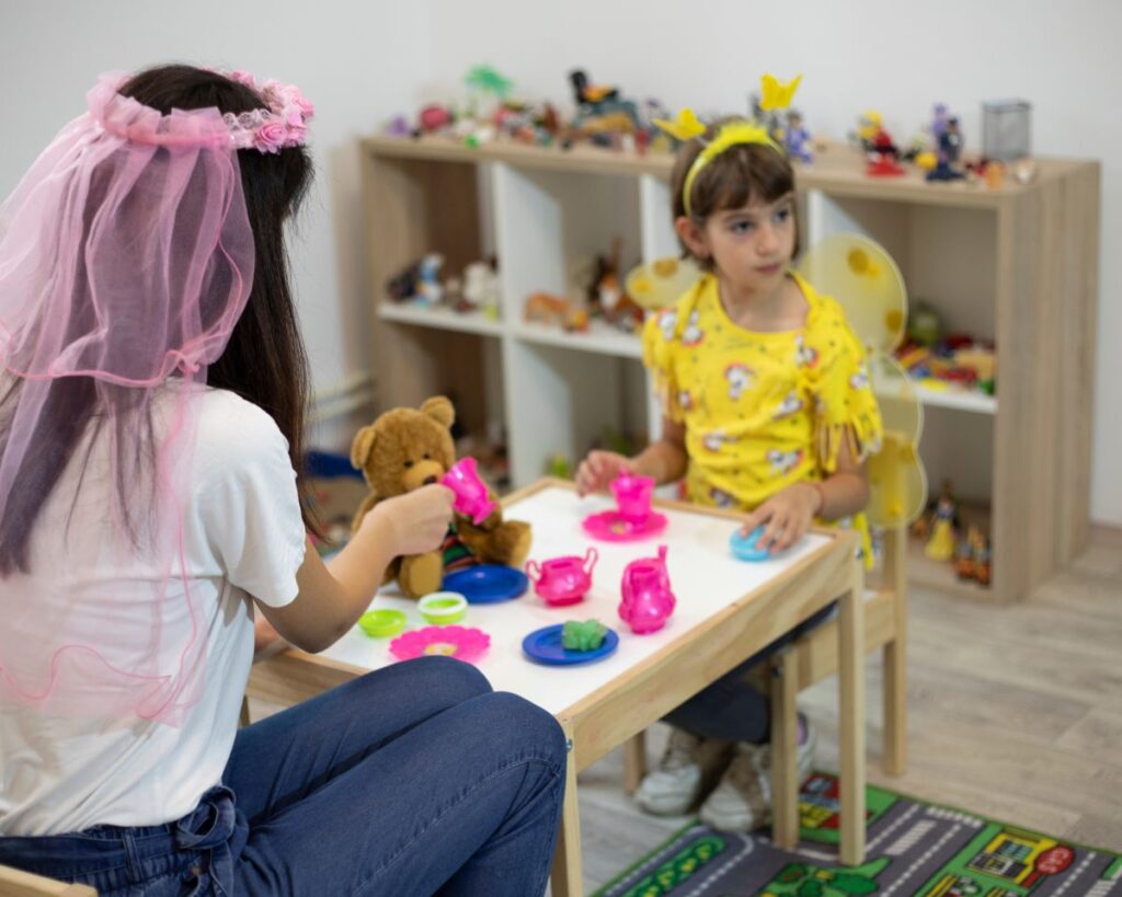 Therapy for Children: Types of therapy that work well for children, what therapy sessions look like, and how parents can be involved. Photo of a young girl in a yellow costume in a playroom, having a tea party with a female therapist.